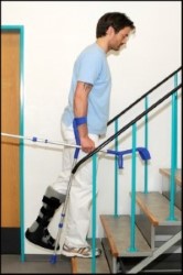 Person ved trappe træder op på trin med rask ben og løfter bandageret ben og stokke op til trin