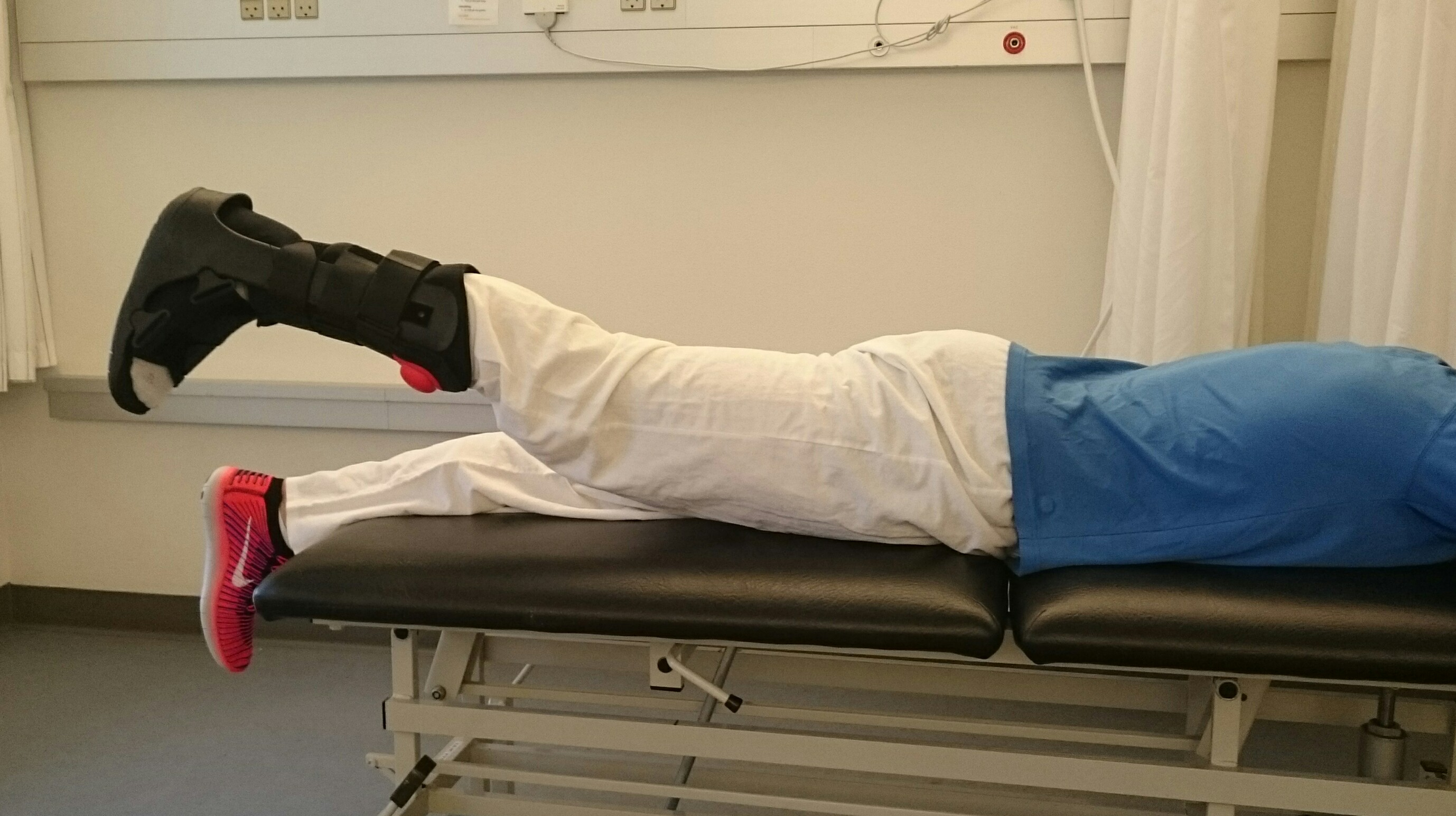Person ligger på maven og løfter et strakt ben med bandage op fra underlaget