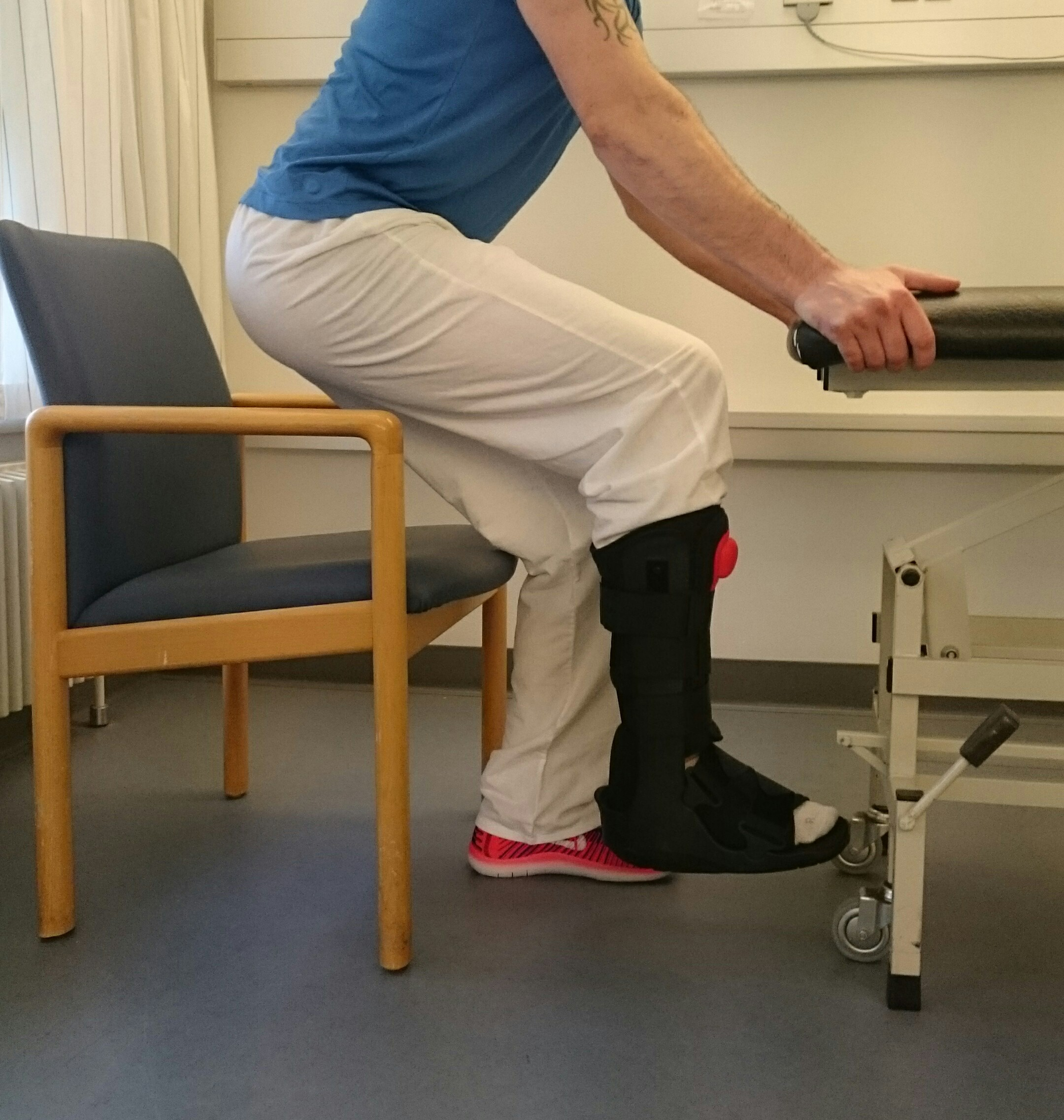 Person står på et ben ved bord og foran en stol. Personen bøjer knæet på standbenet