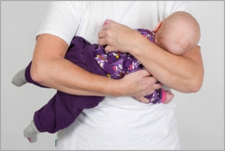 Voksen bærer barn sideliggende mod sin mave og skubber barnets hoved mod venstre.