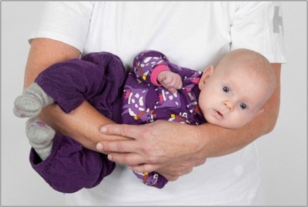 Voksen bærer barn sideliggende med ryggen mod maven og bøjer barnets hoved mod venstre med armen