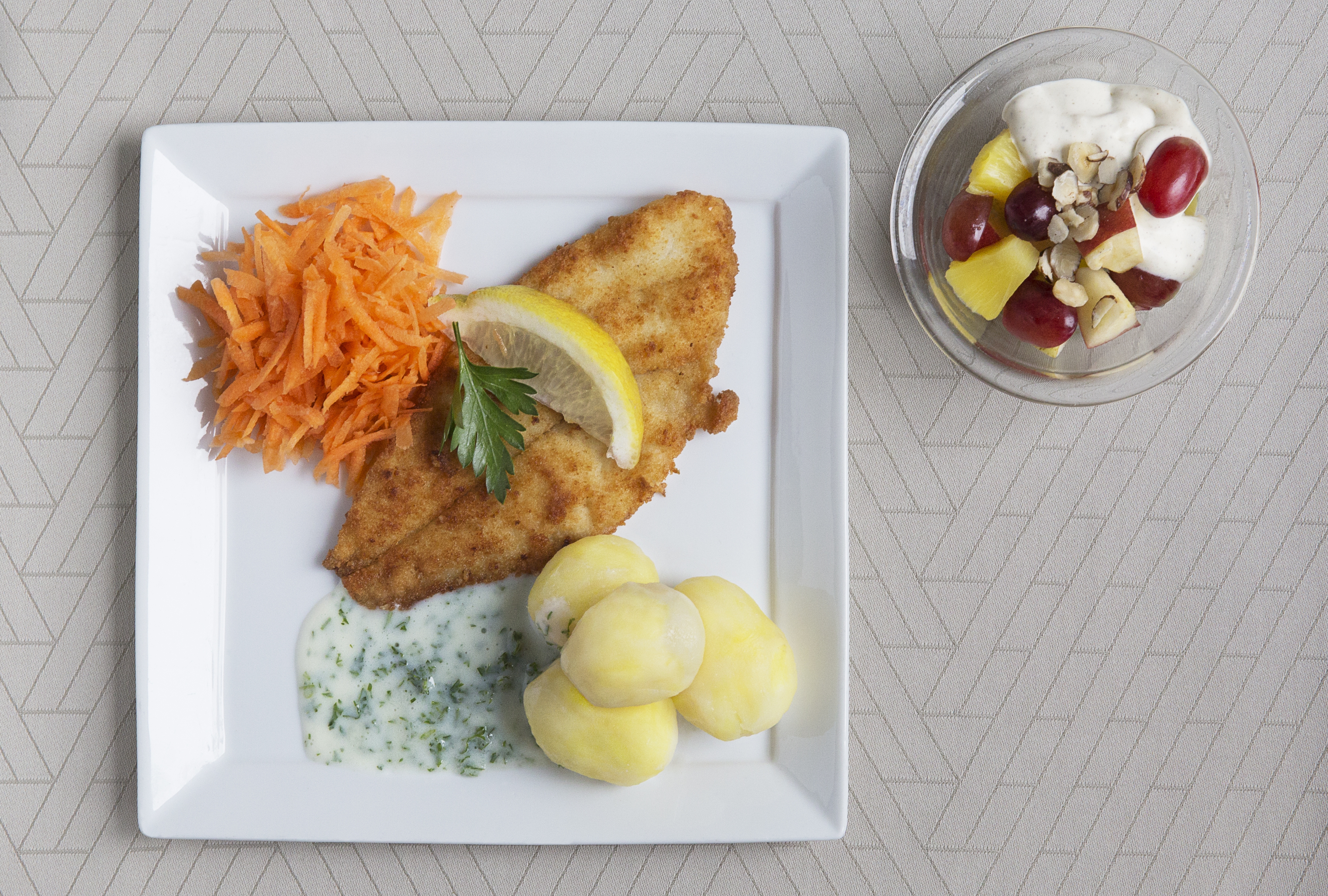 Tallerken med fiskefilet, kartofler, revet gulerod, persillesovs og skål med vindruer, ananas, æblestykker, råcreme og nødder