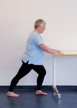 Patienten står op og holder ved et bord for at holde balancen. Venstre ben er bøjet let i knæet, det højre ben er strakt med hælen i gulvet. Fødderne peger lige frem.