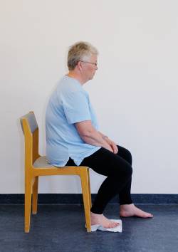 Patienten sidder med rank ryg på kanten af en stol med bare fødder. Under den ene fod ligger en karklud.
