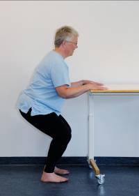 Patienten står med let bøjede knæ foran et bord. Patienten har hænderne placeret på bordet for at holde balancen.